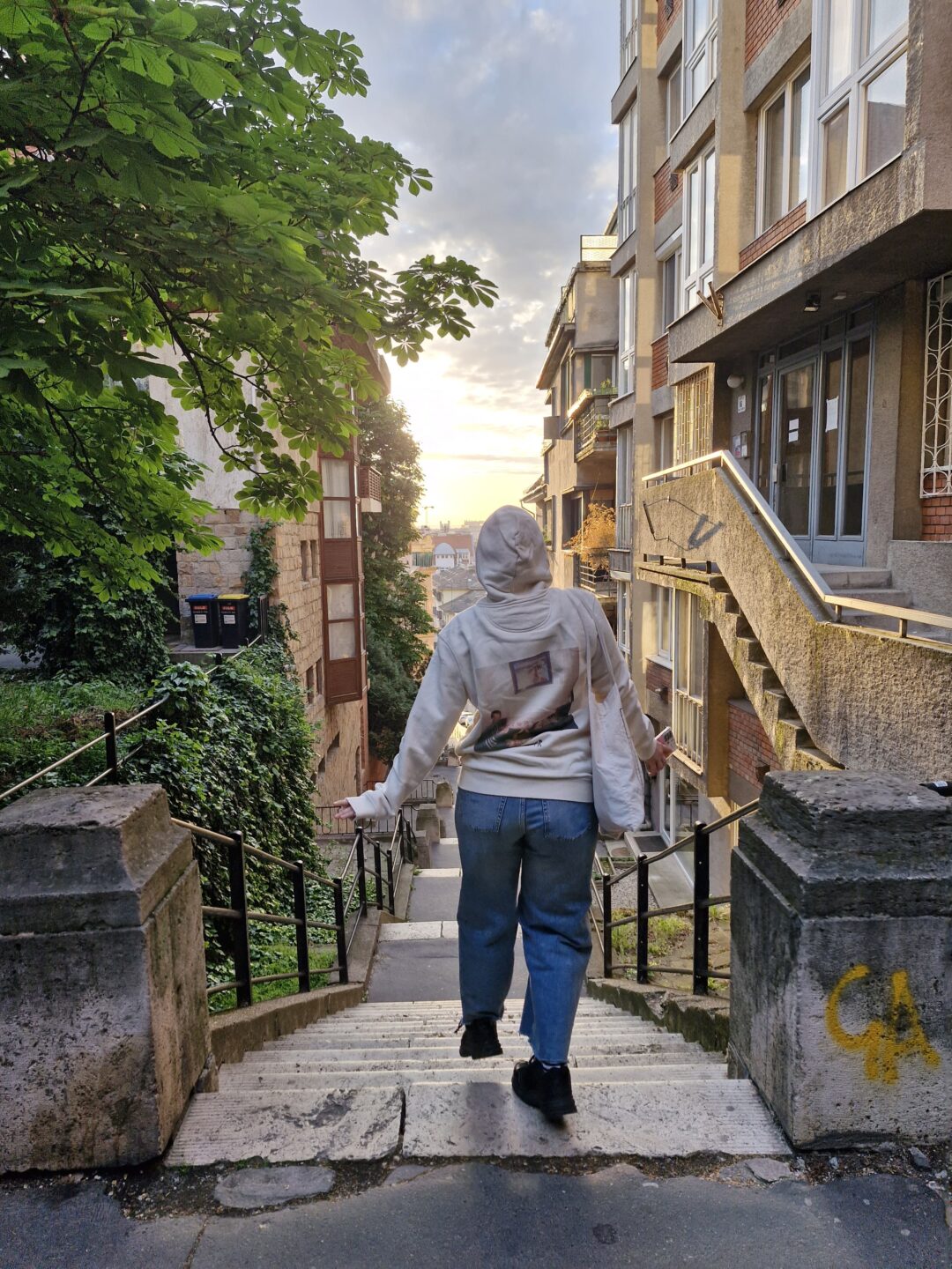 Auf dem Bild sieht man eine Frau, welche auf einer Treppe, am Anfang einer Gasse steht. Im Hintergrund geht die Sonne auf.