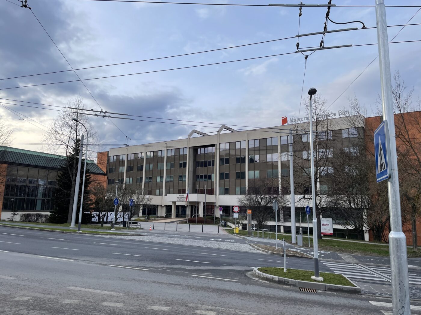 Auf dem Bild ist ein aus Beton bestehendes, weißes Unigebäude zu sehen, davor sieht man die Straße und eine tschechische Flagge