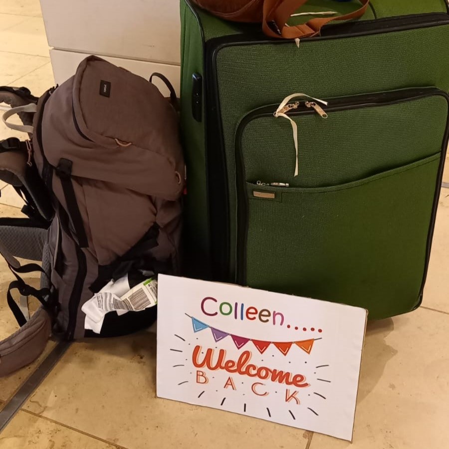 Auf dem Bild sieht man einen Backpacking-Rucksack und einen großen grünen Koffer. Davor steht ein Schild auf welchem Steht "Colleen...Welcome Back".