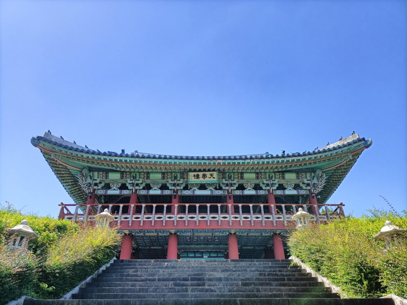 Ein großer rot grüner Tempel steht im Zentrum des Bildes. Eine Treppe führt zu diesem herauf. Der Himmel ist strahlend blau ohne Wolken.