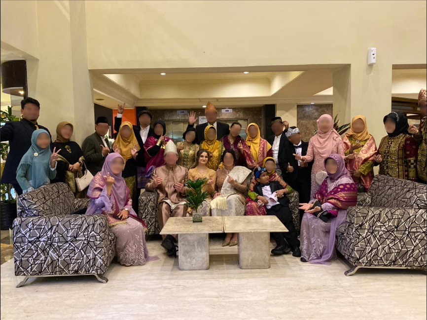 Eine große Gruppe von Menschen in traditioneller indonesischer Kleidung versammelt sich in einer eleganten Lobby. Die Teilnehmer, sowohl Männer als auch Frauen, zeigen eine Vielzahl von Farben und Mustern in ihren Outfits. Die Personen posieren fröhlich für die Kamera, einige sitzen auf Sofas. Die Umgebung ist hell erleuchtet und vermittelt eine festliche Atmosphäre. Im Hintergrund sind Pflanzen und dekorative Elemente sichtbar, die zur Gesamtästhetik des Raumes beitragen.