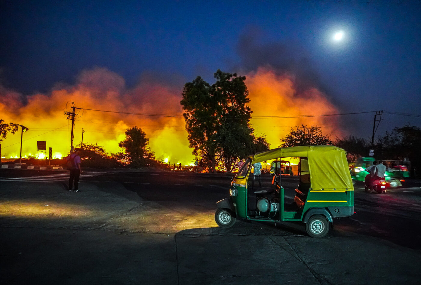 What Stayed After the Culture Shock 27 DSC00337 An auto rickshaw parked on a road at night, with large flames and smoke burning in the background under a bright moon.