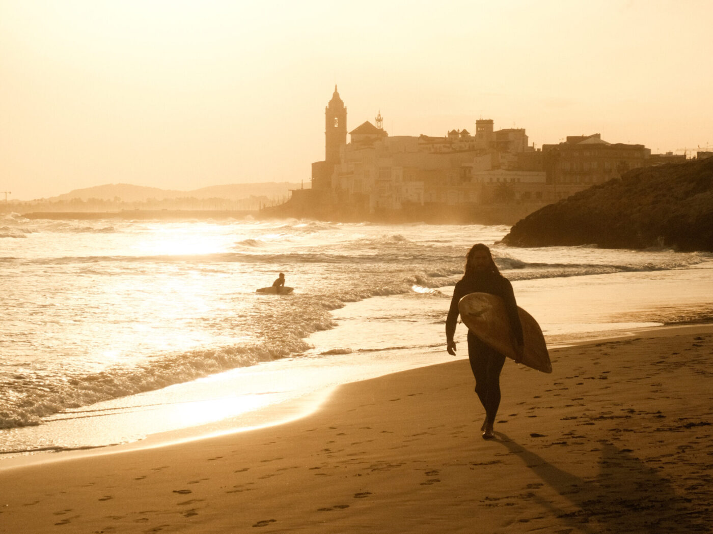 "Living La Vida Loca" in Barcelona 1 DSCF4930 Person carrying a surfboard walking along a beach at sunset with a coastal town in the background.